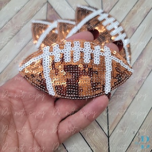 May include: A football-shaped hair bow, covered in bronze sequins, with white accents. The bow is held in a hand, and the design mimics a football. The background is a light wood grain pattern.