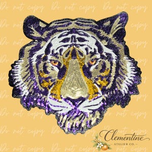 May include: A tiger head patch crafted with purple, gold, white, and black sequins. The tiger features piercing orange eyes, a detailed striped pattern, and a textured finish. The design is symmetrical, showcasing the feline's face with intricate beadwork.