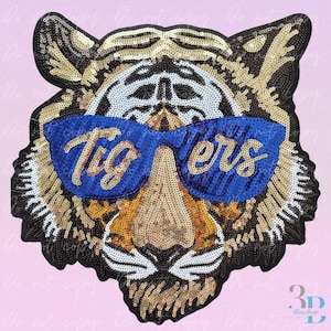 May include: A sequined tiger face patch with blue sunglasses that read "Tigers". The patch features gold, black, white, and tan sequins. The patch is likely for clothing or accessories.