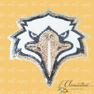 Könnte beinhalten: Ein paillettenbesetzter Adlerkopf-Aufnäher mit weißen, goldenen und schwarzen Details. Der Aufnäher zeigt ein detailliertes Design eines Adlergesichts mit einem goldenen Pailletten-Schnabel und goldenen und schwarzen Pailletten-Akzenten. Der Aufnäher befindet sich auf einem gelben Hintergrund.