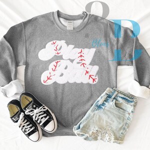 May include: A gray sweatshirt with the words "Play Ball" in a baseball-themed design, paired with denim shorts and black Converse sneakers. The sweatshirt features white lettering with red stitching details, creating a sporty aesthetic.