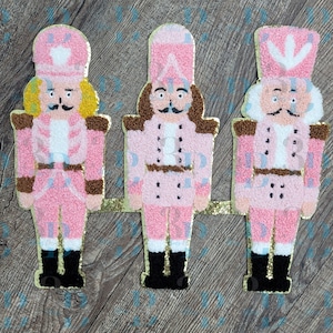 May include: Three nutcracker figures crafted with a textured punch needle finish in shades of pink, white, and brown. Each figure wears a tall hat and uniform, featuring black boots and gold-toned accents, connected by a horizontal gold base.