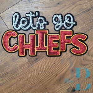 May include: A sequined patch with the words "let's go CHIEFS". The text "let's go" is in a light blue color, and the word "CHIEFS" is in red with a yellow outline. The patch is on a wooden surface.