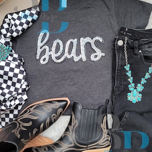 May include: A flat lay featuring a dark gray t-shirt with the word "bears" in silver sequins, paired with black jeans, a turquoise necklace, and black cowboy boots with gold embroidery. A checkered scarf and a cowboy hat are also present.