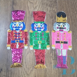 May include: Three sequined nutcracker patches. The left one is magenta and red, the center one is green and red, and the right one is pink and silver with a black hat. Each figure features a tall hat, a uniform jacket, and a belt.