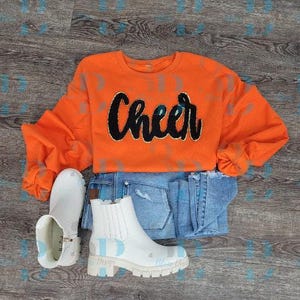 May include: An orange sweatshirt with the word "Cheer" in black script, paired with blue jeans and white ankle boots. The boots have a chunky sole and a decorative design. The outfit is laid out on a wooden surface.