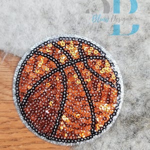 Peut inclure: Un écusson de basket-ball rond avec des paillettes orange et des coutures noires, bordé de perles argentées. L'écusson porte le texte "3D Blues Design" dans le coin supérieur droit.