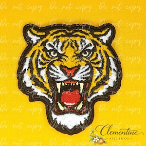 May include: An embroidered patch featuring the head of a tiger with its mouth open, displaying sharp teeth and a red tongue. The tiger has yellow, black, and white fur patterns, outlined in a thick black border against a bright yellow background.