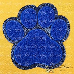 May include: A blue paw print patch featuring a textured, fuzzy center for each pad and a dark, glittery border surrounding the entire shape. The design consists of one large central pad and four smaller rounded pads arranged in a paw pattern.