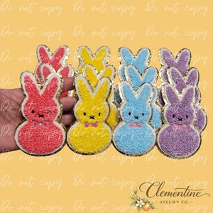 May include: A collection of colorful bunny-shaped patches. Each patch features a different color: red, yellow, blue, and purple. They have gold glitter edging, bow ties, and embroidered facial features. The patches are likely for crafts or embellishments.