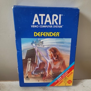 Puede incluir: Una caja azul del videojuego Atari Defender. La caja presenta el logotipo de Atari y el título del juego. La portada muestra una escena futurista con una mujer y una nave espacial. La caja también incluye el texto "COMIC BOOK".