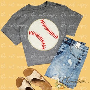 Puede incluir: Una camiseta gris con un diseño de béisbol, combinada con pantalones cortos de mezclilla y sandalias. El gráfico de béisbol es blanco con costuras rojas y un borde dorado. Los pantalones cortos son de color azul claro con detalles desgastados.