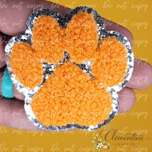 May include: A paw print shaped patch featuring a textured orange fuzzy center surrounded by a shimmering silver glitter border. The design consists of four rounded toe pads and a larger central pad, all outlined in a reflective silver material.