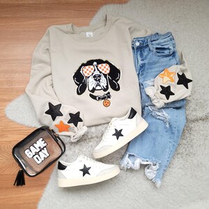 May include: A beige crewneck sweatshirt featuring a graphic of a dog wearing orange and white checkered sunglasses and a collar with a round tag. The sleeves have black and orange star patches. Paired with distressed blue jeans, white sneakers with black star accents, and a clear bag with the text GAME DAY.