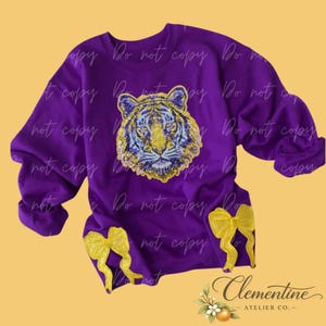 Puede incluir: Una sudadera de manga larga de color morado vibrante con un parche central de cabeza de tigre de lentejuelas en tonos dorados y morados, complementada con dos lazos decorativos de lentejuelas doradas cerca del dobladillo inferior.