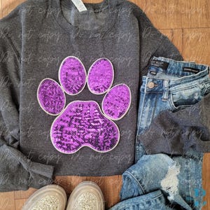 May include: A dark gray sweatshirt with a large purple sequined paw print design. The image also includes a pair of blue jeans, a pair of silver glitter sneakers, and a wooden surface.
