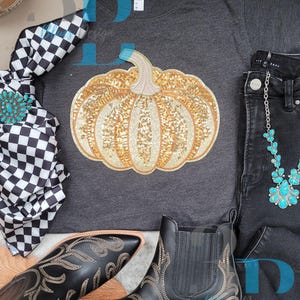 Può includere: Una t-shirt grigio antracite con una grande grafica centrale a forma di zucca decorata con paillettes dorate e arancioni. La maglietta è presentata insieme a jeans neri, una sciarpa a scacchi bianchi e neri con una spilla turchese e stivaletti neri.