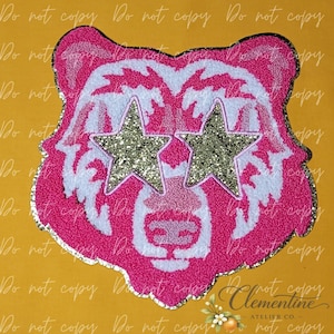 Puede incluir: Un parche de chenilla con forma de cabeza de oso en rosa brillante y blanco. El oso presenta dos grandes ojos en forma de estrella con purpurina dorada y un borde de purpurina plateada que perfila toda la forma de la cabeza del oso.