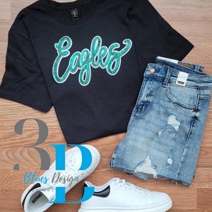 May include: A black t-shirt with the word "Eagles" in teal glitter script, paired with distressed denim shorts and white trainers. The trainers have black accents. A casual outfit.