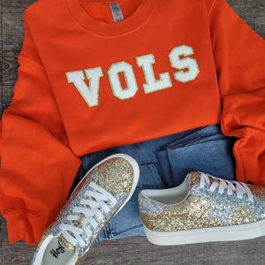 May include: A bright orange crewneck sweatshirt with the word VOLS in white block letters with gold glitter trim, paired with blue denim jeans and a pair of low-top trainers featuring a gold and silver glitter pattern and white laces.