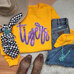 May include: A flat lay arrangement featuring a yellow sweatshirt with the word "Tigers" in purple sequins, alongside blue denim jeans, brown cowboy boots, a turquoise necklace, a hat, and a black and white checkered scarf.