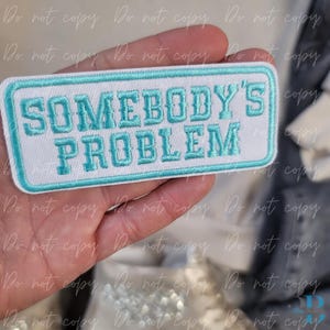 Puede incluir: Un parche rectangular blanco bordado con las palabras "SOMEBODY'S PROBLEM" en letras turquesas. El parche tiene un borde turquesa y se sostiene en una mano.