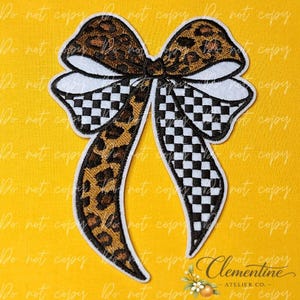 May include: An embroidered patch shaped like a bow featuring a combination of brown and black leopard print fabric and a black and white chequered pattern. The bow has two loops and two long, trailing tails with a white border outline.