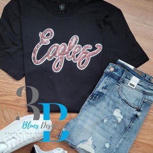 May include: Black t-shirt with the word "Eagles" in pink glitter script, paired with distressed denim shorts and white trainers. The shirt is a crew neck style.