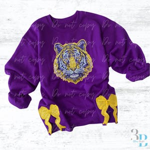 May include: A purple sweatshirt with a sequined tiger face in gold and blue. Two gold bow appliques are sewn on the sleeves. The words "Do not copy" are printed repeatedly across the sweatshirt.
