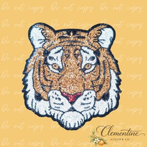 May include: A sequined tiger head patch with a black outline, white accents, and a red nose. The tiger's face is made of gold sequins. The background is a light yellow color. The text "Clementine ATELIER CO." is in the bottom right corner.