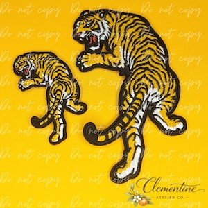 May include: Two embroidered tiger patches in different sizes, featuring a leaping tiger design with yellow fur, black stripes, and white accents. Each tiger is depicted in a dynamic pouncing pose with an open mouth showing red details.