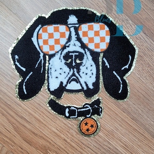 May include: Embroidered patch featuring a dog with sunglasses. The dog is black and white with orange and white checkered sunglasses. The patch has a gold glitter border and a collar with a charm.