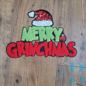 Può includere: Una toppa decorativa con un cappello da Babbo Natale in paillettes rosse e bianche sopra la scritta MERRY GRINCHMAS. La parola MERRY è in ciniglia verde, mentre la parola GRINCHMAS è in ciniglia rossa con un piccolo ornamento rosso a paillettes.