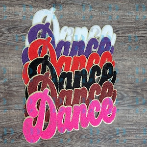 Könnte beinhalten: Ein Stapel bunter, gestickter "Dance"-Patches in verschiedenen Farben, darunter Weiß, Lila, Rot, Schwarz und Pink. Jeder Patch hat einen goldenen Rand und eine strukturierte Oberfläche, ideal für Kleidung oder Bastelarbeiten.