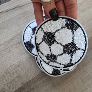 Könnte beinhalten: Kreisförmige Aufnäher mit einem Fußball-Design. Die Aufnäher sind weiß mit schwarzen Fünfecken und Sechsecken und mit Pailletten bedeckt. Die Aufnäher sind auf einer hellen Holzoberfläche gestapelt.