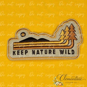 May include: An embroidered patch with a light gray background featuring a black mountain silhouette, a small sun, and three stylized pine trees in shades of brown and gold. The text KEEP NATURE WILD is stitched in black at the bottom of the patch.