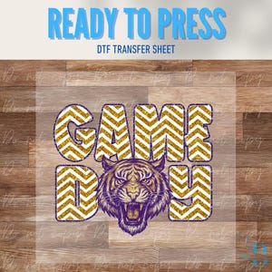 May include: A DTF transfer sheet with the words "GAME DAY" in gold chevron pattern letters. A purple and gold tiger graphic is centered below the text. The words "READY TO PRESS" and "DTF TRANSFER SHEET" are at the top.