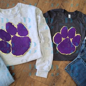 May include: A light gray sweatshirt and a dark gray t-shirt, both featuring a large purple paw print with gold trim. The items are displayed with a pair of blue jeans on a wooden surface. The sweatshirt has long sleeves.