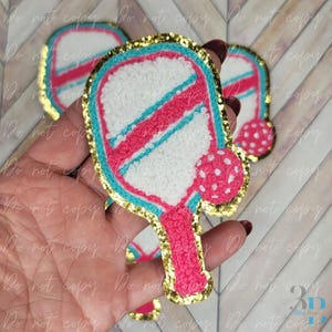 May include: A colorful pickleball paddle and ball design with a gold glitter border. The paddle features a white center with pink and teal stripes, and a pink handle. The ball is pink with white polka dots. The design is on a white background.