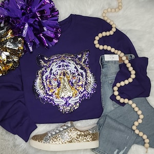 Op de afbeelding: Een paars sweatshirt met lange mouwen en een grote lovertjesapplicatie van een tijgerkop in paars, goud en zilver. De outfit bevat een lichtblauwe spijkerbroek met een zichtbaar label maat 3/26, gouden glittersneakers en een paars-gouden pompon.