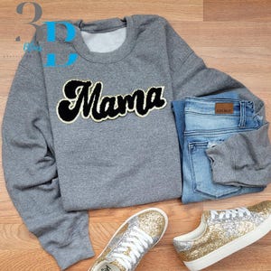 Pode incluir: Um moletom cinza com a palavra "Mama" em um patch preto e dourado, combinado com jeans azuis e tênis dourados com glitter. O conjunto é apresentado em uma superfície de madeira, mostrando um estilo casual.