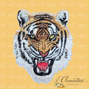 Può includere: Toppa ricamata con testa di tigre ruggente. La toppa presenta un design dettagliato con colori arancione, nero e bianco e una bocca rossa. Il testo "Clementine - ATELIER CO." è in basso.
