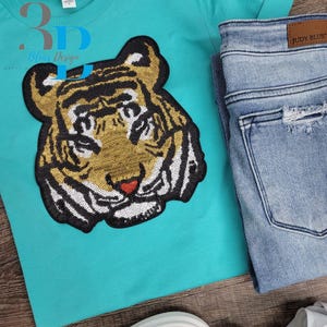 Puede incluir: Una camiseta turquesa con un diseño de cara de tigre con lentejuelas. El tigre tiene detalles dorados, negros y blancos con una nariz roja en forma de corazón. Vaqueros de mezclilla azul claro doblados junto a la camisa. El texto "3D Blue Design" es visible.