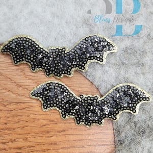 May include: Two bat-shaped patches featuring black sequins covering the wings and body, outlined with a gold-colored border. The patches are positioned against a textured gray and wood-grain surface. The text 3D Blues Designs is visible in the upper right corner.