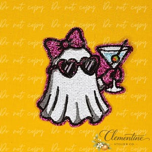 May include: An embroidered patch featuring a white ghost wearing a pink glittery bow and black heart-shaped sunglasses. The ghost holds a martini glass with a green olive. The entire design is outlined in a vibrant pink glittery border.