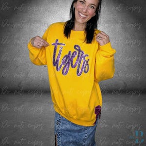 May include: A yellow sweatshirt with the word "tigers" in purple glitter lettering. The sweatshirt has a purple bow detail on the lower right side. The person is wearing a denim skirt.