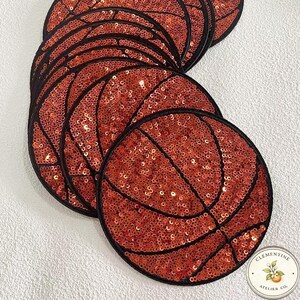 May include: A collection of sequined basketball patches. Each patch is round, with a black outline and a design mimicking a basketball. The patches are covered in orange sequins, with black lines to represent the basketball's panels. The patches are arranged on a white surface.