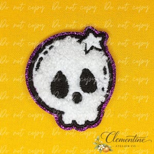 Puede incluir: Un parche bordado de calavera blanca con una superficie texturizada y difusa, que presenta grandes cuencas oculares negras y una pequeña nariz negra. La calavera está delineada con un borde de purpurina morado oscuro y tiene una pequeña estrella blanca.