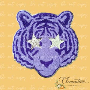 May include: A tufted rug shaped like a tiger head in shades of purple and lavender. The tiger features dark purple stripes, a light purple muzzle, and two silver glittery star-shaped patches covering the eye area, creating a distinct graphic design.