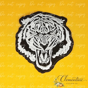 May include: An embroidered patch featuring the head of a tiger with its mouth open in a roar. The design uses white thread for the tiger's fur and facial features against a solid black background, creating a high-contrast graphic illustration.